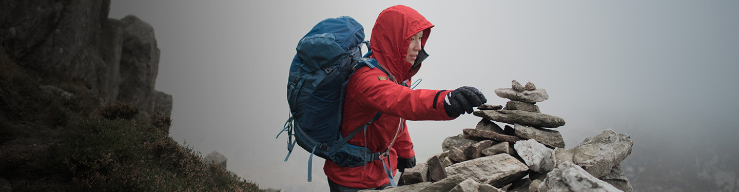 A woman in a red waterproof jacket and a rucksack places a stone on a cairn.